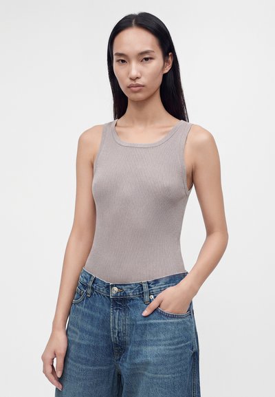 Mujer con cabello largo y negro, que lleva una camiseta de canalé beige sin mangas y vaqueros azules sueltos con una mano en el bolsillo, sobre un fondo liso.