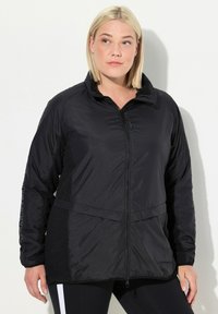 Veste isolante noire avec fermeture éclair à l'avant, col haut et panneaux latéraux texturés. Présente une taille cintrée et est fabriquée en tissu léger.