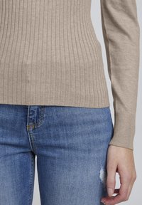 Beige ribbat stickad tröja med långa ärmar, kombinerad med blå denimjeans med en slitning vid knät.