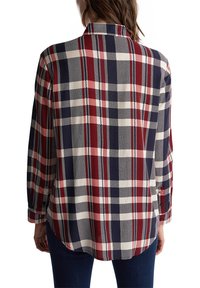 Donna che indossa una camicia a maniche lunghe a quadri rossi, blu navy e bianchi, in piedi con le spalle rivolte verso la macchina fotografica.