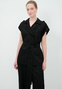 Gilet noir à double boutonnage avec des manches courtes, ceinture à nouer et accents de boutons. Porté sur un pantalon noir à jambes larges, tissu au toucher lisse.