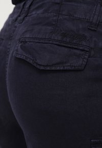 Detailní záběr na zadní kapsu tmavě modrých kalhot s klopou a vyšitým názvem značky "Kapapapi" nad kapesními otvory.