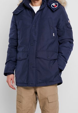 Homme portant une veste d’hiver bleu marine avec une capuche bordée de fourrure et un pantalon cargo beige sur un fond uni.