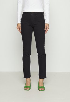 Donna che indossa jeans neri aderenti, top bianco a maniche lunghe e sandali verdi con tacco aperto, in piedi davanti a uno sfondo chiaro e uniforme.