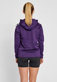 Felpa sportiva viola con cappuccio e maniche lunghe. Presenta dettagli riflettenti sulle spalle. Abbinata a pantaloni sportivi neri.