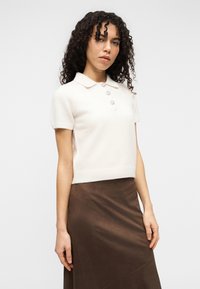 Κοντομάνικο, ελεφαντόδοντο πόλο τοπ με γιακά και τρία διακοσμητικά κουμπιά. Μαλακό, υφασμάτινο υλικό σε συνδυασμό με μακριά, καφέ φούστα.