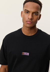 Νέος άνδρας με κοντά σγουρά μαλλιά, φοράει μαύρο T-shirt με μικρό ορθογώνιο λογότυπο "LES DEUX" στο στήθος, μπροστά σε απλό φόντο.