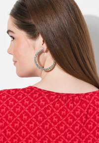 Top rojo con un patrón y motivos sutiles de ciervo. Pendiente grande de aro plateado adornado con cristales claros. Cabello marrón liso y lacio.