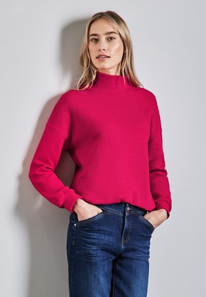 Fuchsia Strickpullover mit hohem Kragen und strukturiertem Muster; kombiniert mit dunkelblauen Jeans, die Hände in den Taschen, vor einem hellen Hintergrund.