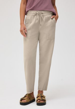 Person iført beige snøre bukser med laplommer og flossede kanter, brune platformsandaler, stående mod en ensfarvet baggrund.