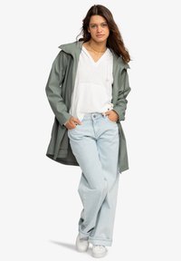 Chaqueta larga impermeable verde sobre una sudadera blanca, combinada con jeans de pierna ancha azul claro y zapatillas blancas. Pose natural y relajada.