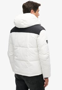 Gevoerde witte en zwarte winterjas met een grote capuchon, voorzien van een gladde textuur en logodetail op de schouder, gezien van achteren.