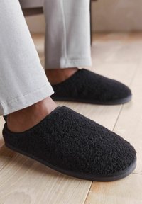 Musta, pörröinen tossu, joka on valmistettu pehmeästä kankaasta, pyöreällä kärjellä, pehmeällä sisäpuolella ja tasaisella kumipohjalla, käytössä vaaleanharmaiden housujen kanssa.