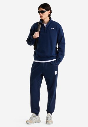 Jeune personne portant un survêtement bleu marine North Face, des baskets blanches, des lunettes de soleil noires et portant un sac à dos sur une épaule.