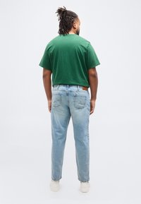 Άνδρας με κότσους dreadlocks, φοράει πράσινο μπλουζάκι, ανοιχτόχρωμο τζιν και λευκά αθλητικά παπούτσια, στέκεται με την πλάτη προς τα εμπρός σε απλό λευκό φόντο.