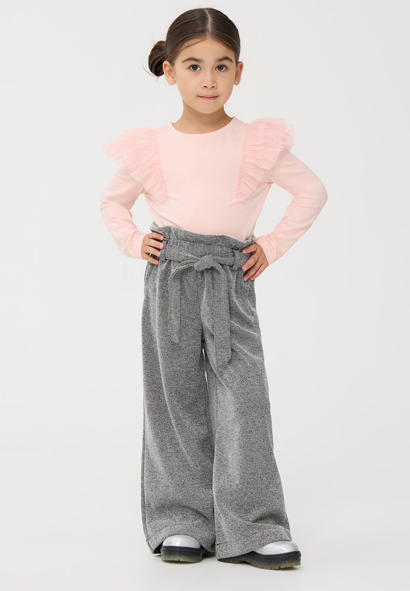Maglietta lunga a maniche corte di colore rosa chiaro con dettagli arricciati e pantaloni grigi a gamba larga con cintura legata, abbinati a scarpe lucide nere e argentate.