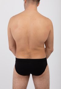 Slips da nuoto neri indossati, con una superficie liscia. La persona è mostrata di spalle, esponendo un torso e gambe nudi su uno sfondo bianco.