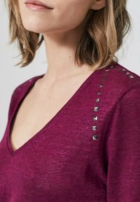 V-ringad, kortärmad topp i djup magentafärgad stickad kvalitet, med metallnitar längs axelsömmen för detalj och textur.