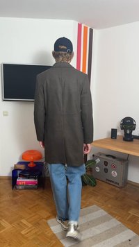 Lange zwarte jas met een rechte snit en een gestructureerde stof, gecombineerd met lichtblauwe jeans en witte sneakers. Achteraanzicht toont een minimalistisch ontwerp.