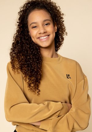 Jonge vrouw met krullend haar, gekleed in een mosterdgele trui, glimlachend met armen gekruist tegen een beige achtergrond.