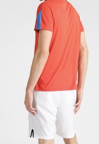 Röd tränings T-shirt med blå detaljer, korta ärmar och avslappnad passform; kombinerad med vita shorts med en sidoklyvning.