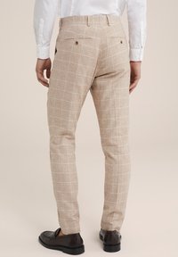 Béžové kostkované kalhoty s texturovanou tkaninou, se subtílním mřížkovým vzorem a dvěma zadními kapsami se zapínáním na knoflíky. V kombinaci s černými botami.