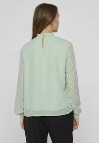 Light green blouse with a textured zigzag pattern, long sleeves, and a ruffled neckline. Features a keyhole back opening and elastic cuffs.