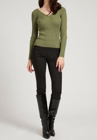 Olive green ribbed long-sleeve top with a V-neck, paired with black fitted pants and knee-high black crocodile-textured boots.
