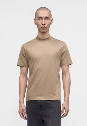 Homme au crâne rasé portant une chemise beige unie à manches courtes et un pantalon foncé, regardant devant avec une expression neutre.