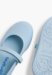Zapatillas de lona azul claro con punta redondeada, que cuentan con una correa con hebilla. La suela tiene texturas con patrones de flores y corazones.