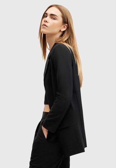 Blazer noir à la coupe ajustée, avec des manches longues, une poche unique et un design cropped. Texture lisse et style minimaliste.