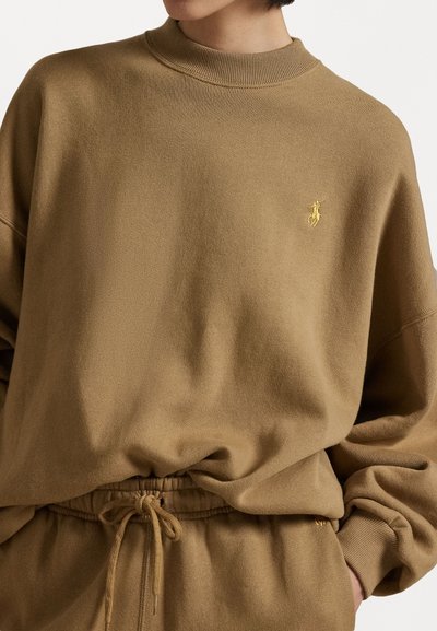 Brown sweatshirt with a high neckline, elastic waistband, and a small gold logo on the chest. Soft texture with a relaxed fit.