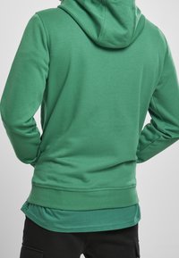 Grüner Hoodie mit weicher Textur, ausgestattet mit einer Kängurutasche und verstellbarer Kapuze mit Kordelzug. Der Saum ist hinten etwas länger.