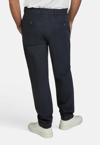 Donkerblauwe broek van een gestructureerde stof, met aan de achterkant twee pockets met metalen knopen, gecombineerd met witte sneakers.