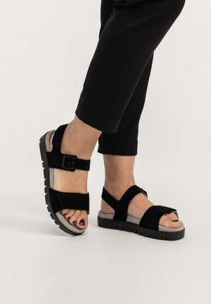 Füße, die schwarze Sandalen mit offenen Zehen, dicken Riemen und Schnallen tragen, kombiniert mit schwarzen, gekürzten Hosen vor einem einfarbigen hellen Hintergrund.