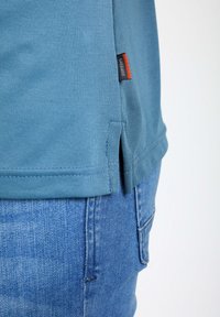 Hellblaues Baumwoll-T-Shirt mit strukturiertem Finish, ausgestattet mit einem seitlichen Schlitze und einem kleinen schwarz-roten Etikett an der unteren linken Seite.