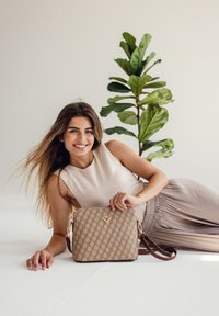 Beige kabelka se vzorem a zlatými detaily, s nastavitelným hnědým popruhem. Hladký povrch s obdélníkovým tvarem, umístěná na světlém pozadí.