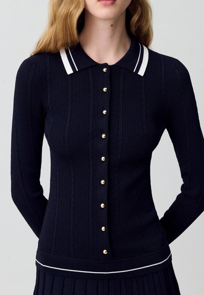 Donna che indossa un cardigan aderente blu navy con bottoni dorati e un colletto con strisce bianche, abbinato a una gonna plissettata coordinata.