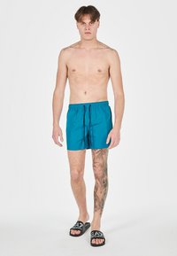 Pantaloni da bagno turchesi con vita elastica e cordino, caratterizzati da una texture liscia. Indossati con ciabatte nere. Il modello ha dei tatuaggi.