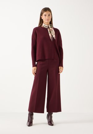 Vrouw draagt een bordeauxrode trui en wijde broek met bijpassende bordeauxrode enkellaarsjes en een bloemenprint sjaal om haar hals.