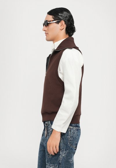 Brauner ärmelloser Pullover mit Reißverschluss, über einem weißen Langarmshirt getragen. Das Model trägt dunkle Sonnenbrillen und gemusterte blaue Jeans.