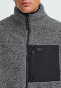 Gray fleece jacket featuring a half-zip design, black fabric pocket with zipper, and a textured surface. Branding tag near the zip.