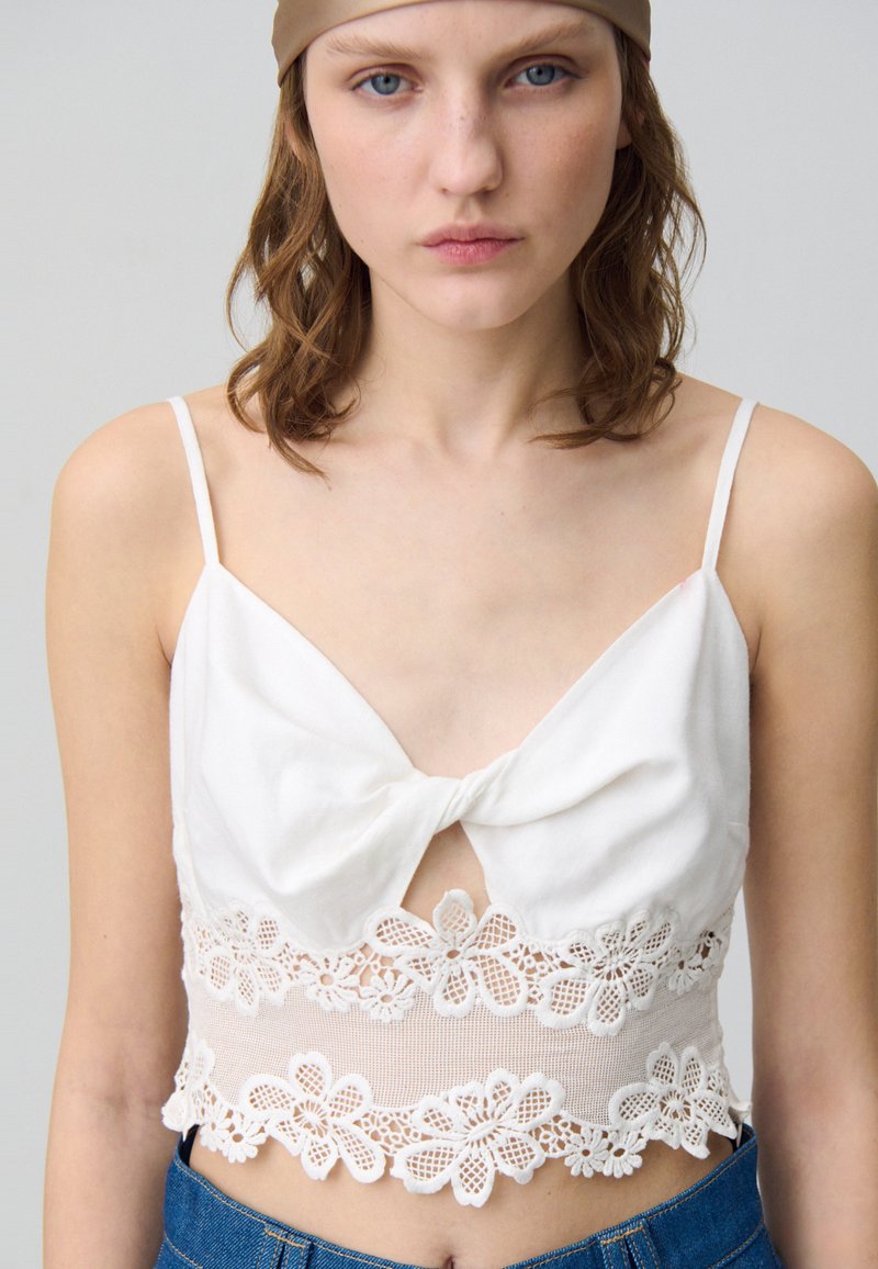 Top blanc court avec un nœud à l'avant, présentant une finition en dentelle avec des motifs floraux. Bretelles spaghetti ajustables et tissu texturé.