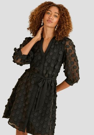 Black dress made of sheer fabric with circular textures, 3/4 sleeves, a V-neck, and a belted waist. Features buttons and lace accents.