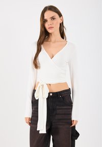 Top bianco avvolgente con maniche lunghe, scollo a V e dettaglio con nodo laterale, abbinato a jeans in denim scuro con vita alta e lievi sbiadimenti.