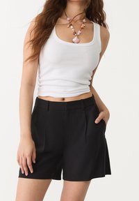 Černé šortky s přizpůsobeným designem, bočními kapsami a jemnými předními sklady. Kombinované s bílým žebrovaným topem a dekorativním náhrdelníkem.