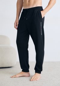 Schwarze Jogginghose mit elastischem Bund, Seitentaschen und einem weißen Logo-Druck am linken Bein. Weich, lockerer Schnitt mit gerippten Bündchen.