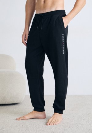 Black sweatpants with an elastic waistband, side pockets, and a white logo print on the left leg. Soft, relaxed fit with ribbed cuffs.