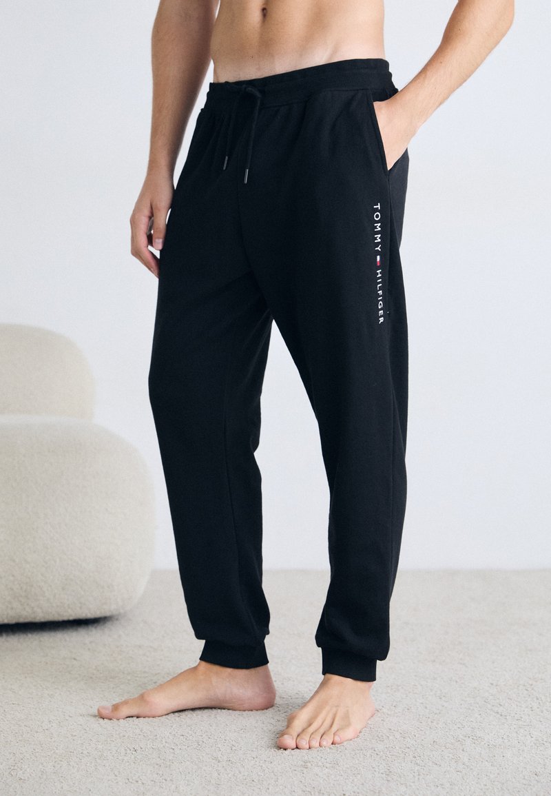 Schwarze Jogginghose mit elastischem Bund, Seitentaschen und einem weißen Logo-Druck am linken Bein. Weich, lockerer Schnitt mit gerippten Bündchen.