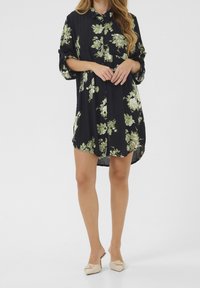 Sort blomsterkjole lavet af let stof med grønne blomsterprint, knaplukning foran og opkrogede ærmer. Fremhævet med en buet kant.